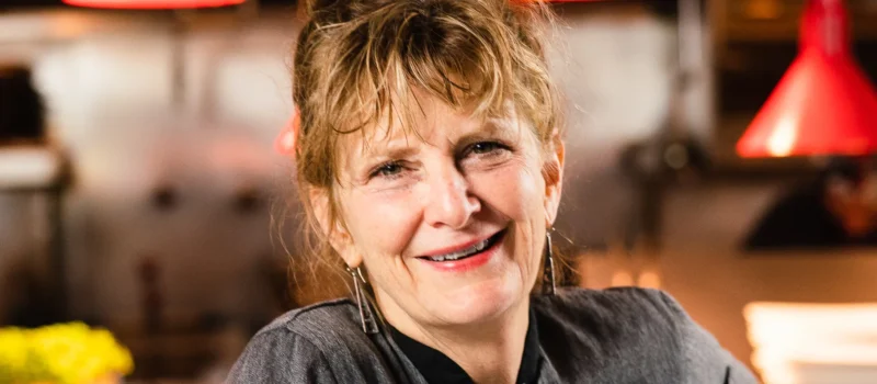 Headshot of chef Deb Paquette, smiling, with red restaurant pendant lights in the background